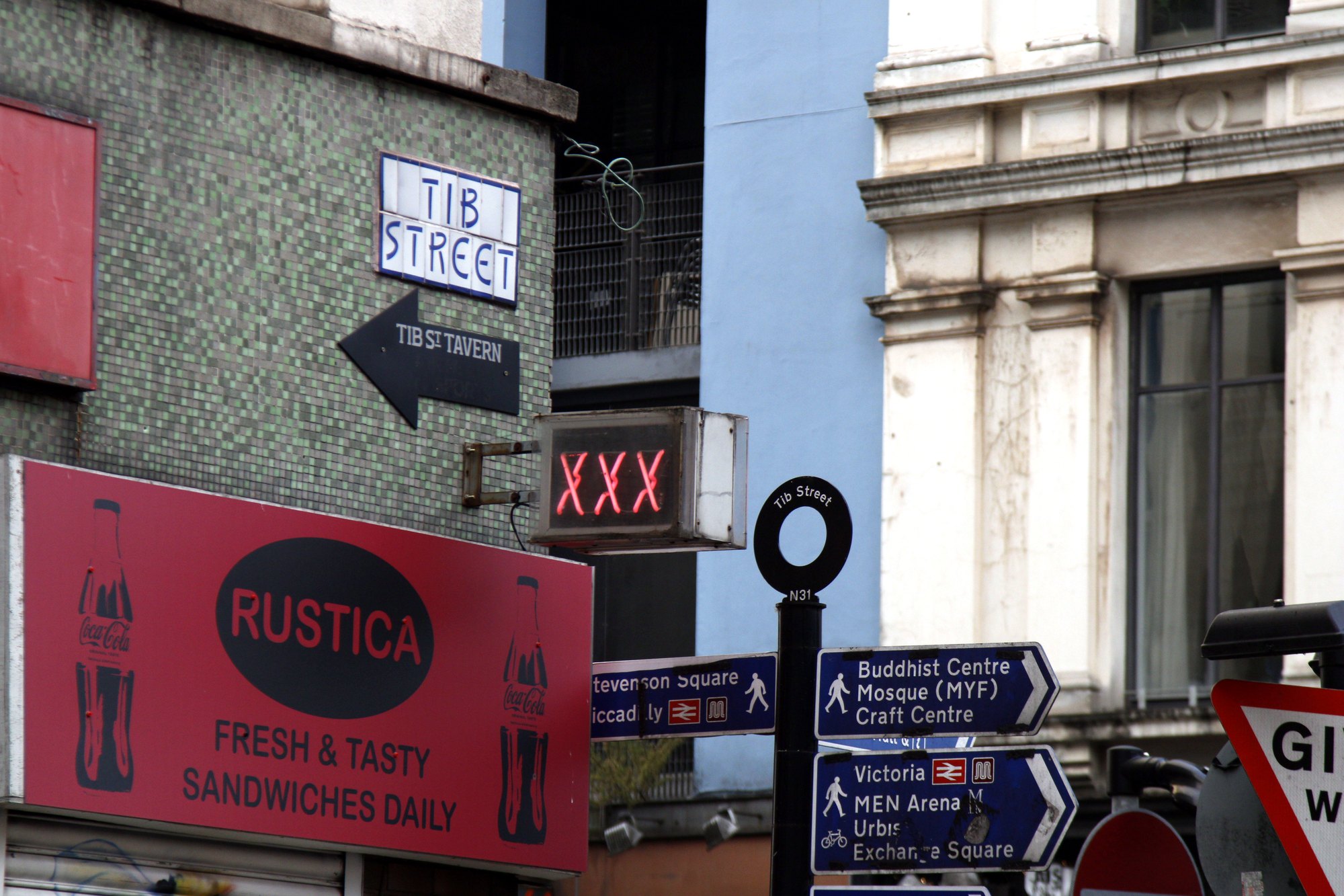 Tib Street sign pointing over a red Rustica sandwich shop sign among Manchester street signs